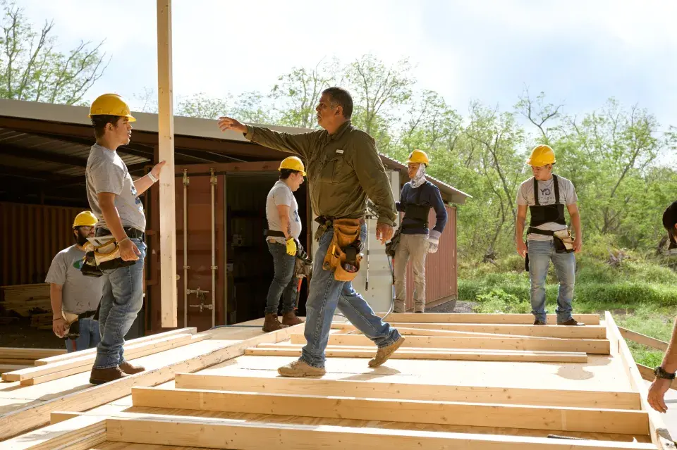 Construction worker stands on the frame of a house with workers in hardhats in the background