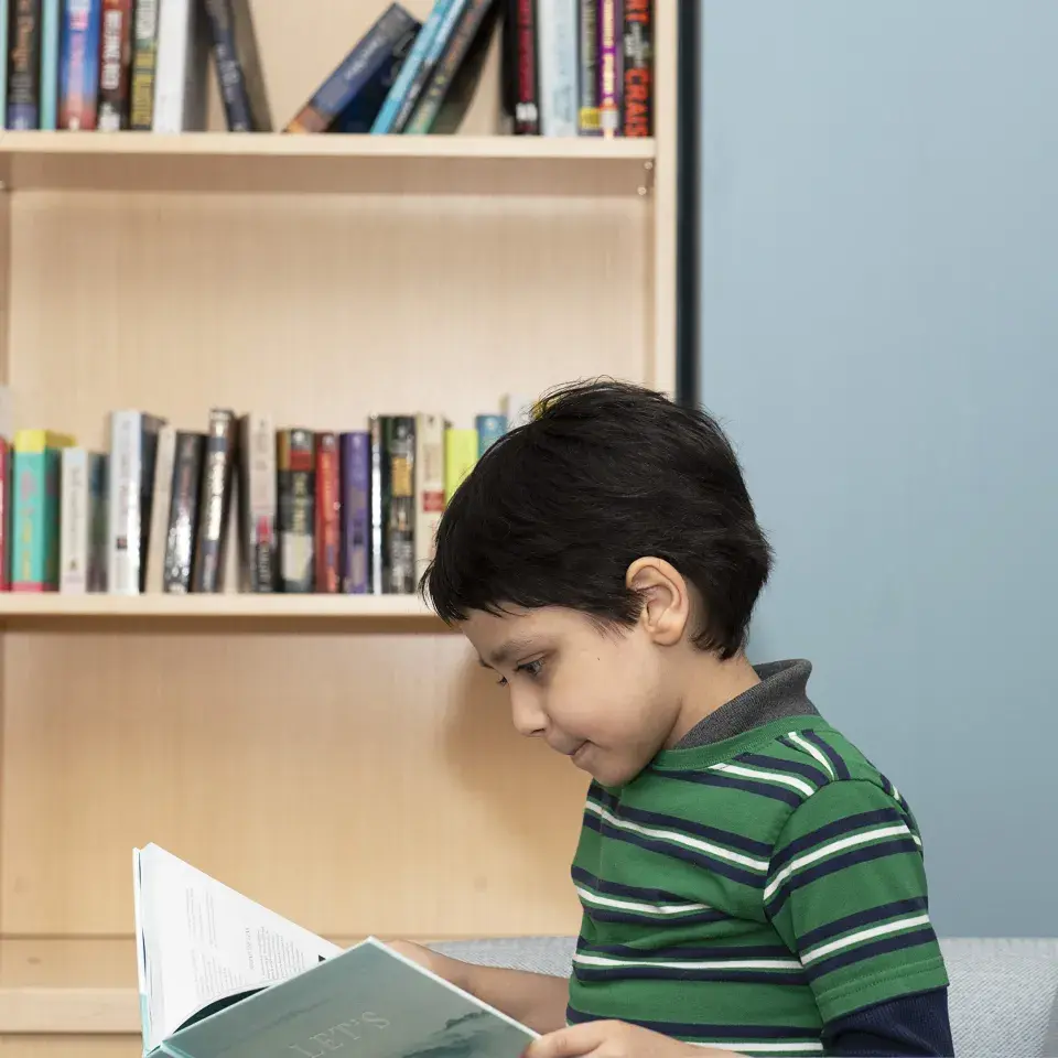 Child sits reading a book with a bookshelf and blue wall in the background