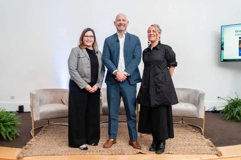 Three people standing on stage with beige velvet chairs in the background
