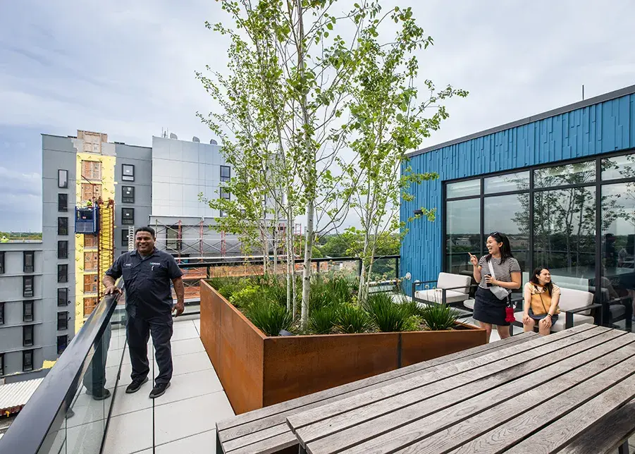 A man poses at a balcony railing, as a woman looks on next to a planter and another woman is seated on a bench. A wooden table is in the foreground and buildings of varying heights in the background