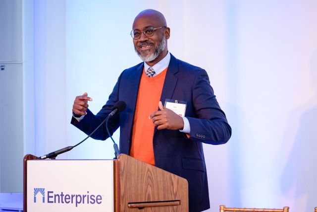 Man with wire glasses and a salt-and-pepper beard wearing a bright orange sweater vest and a blue suit jacket stands speaking at a podium.