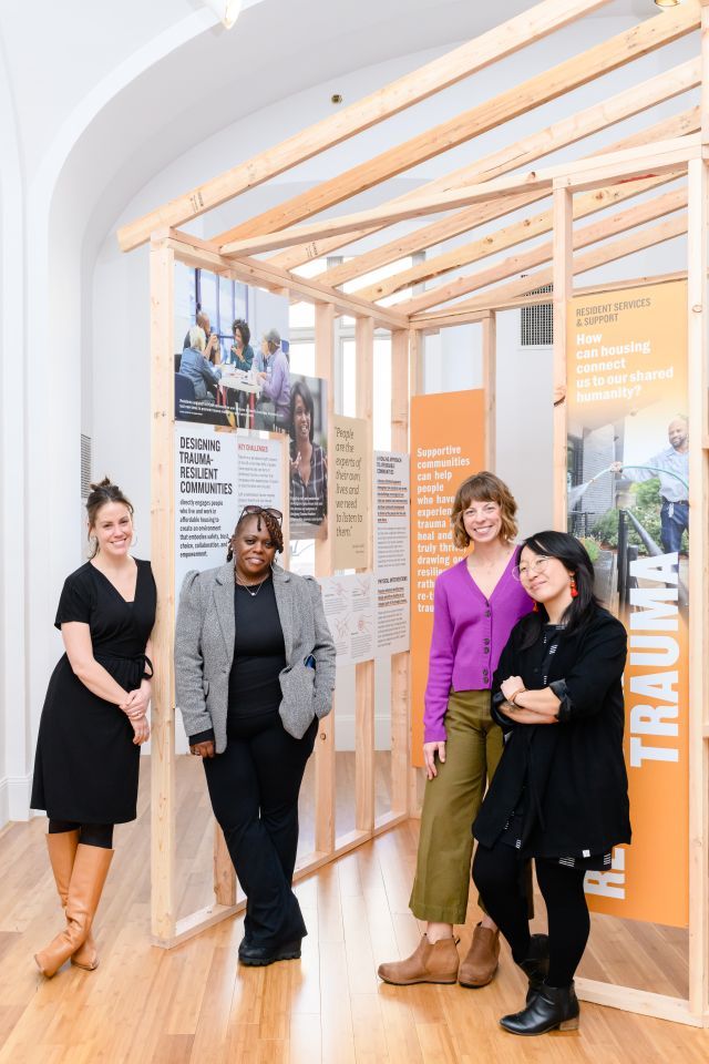 Four women stand in front of a house-like structure featuring photography and text panels.