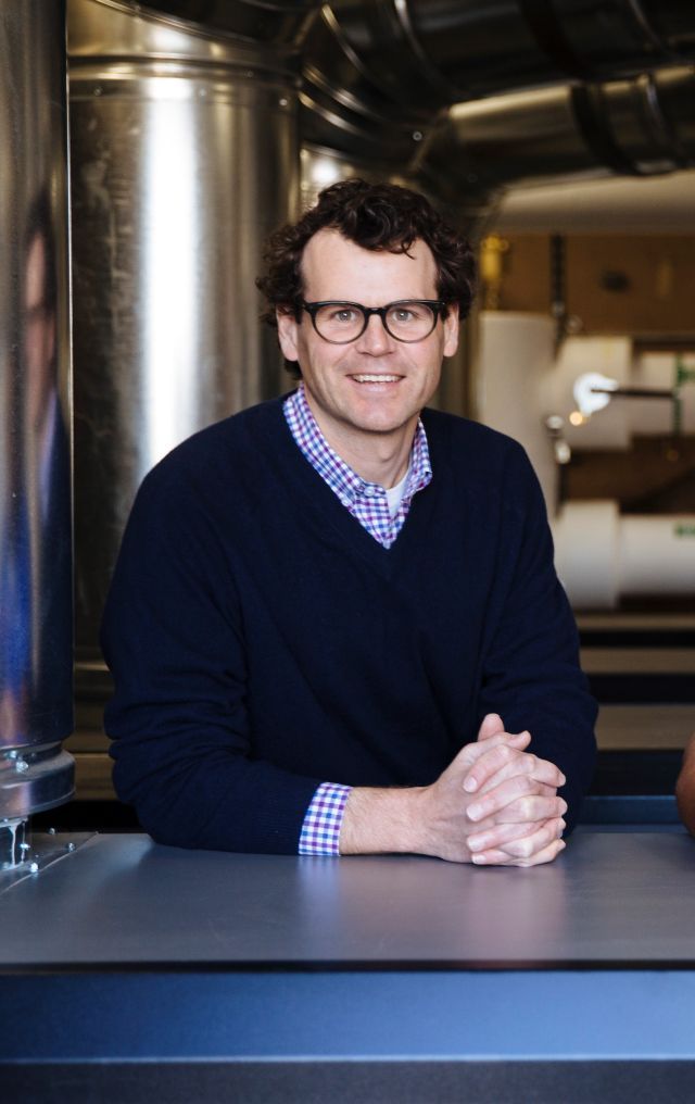 Man with brown glasses and blue v-neck sweater with a blue check pattern shirt with a tall silver cylinder and pipes in the background.