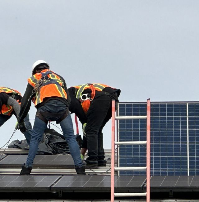 Workers wearing construction hats and orange safety vests install solar panels on a rooftop.