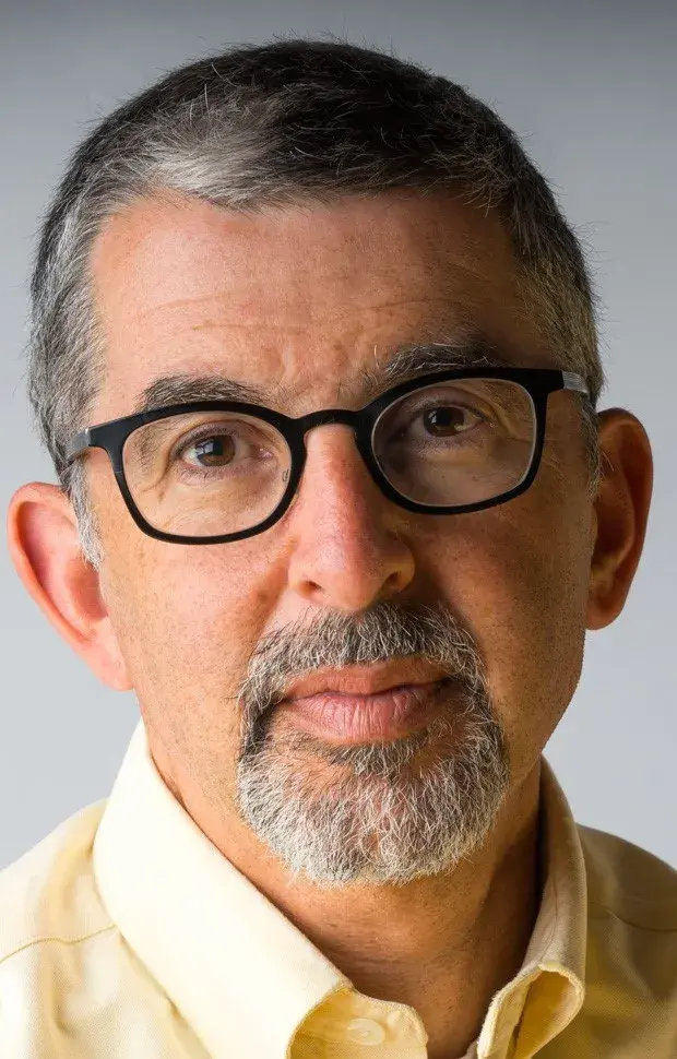 Person with short salt-and-pepper hair wearing black glasses and a yellow collared shirt.