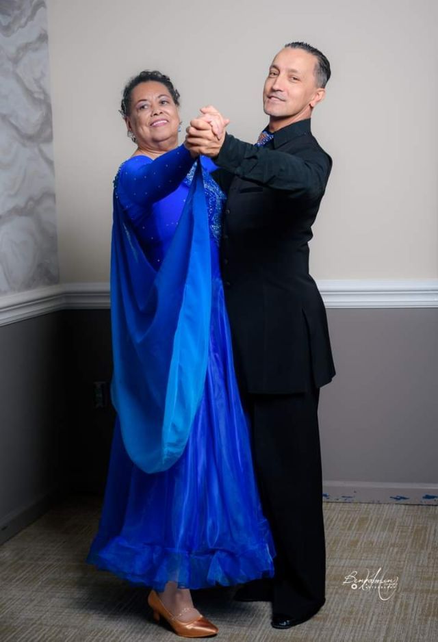 Two people strike a dance pose; one wears blue ball gown and one a dark suit