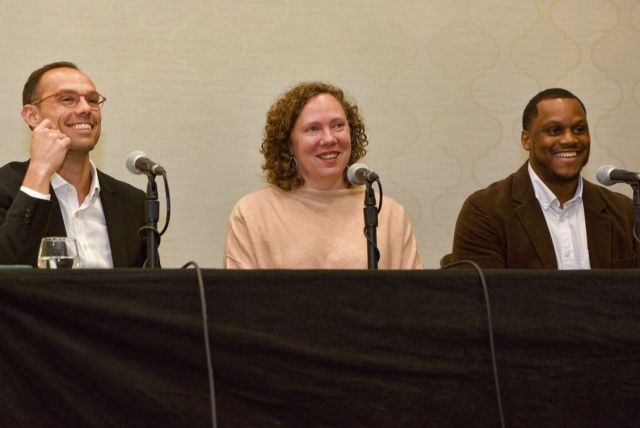 Three people smiling with mics in front of them seated at a table with a dark tablecloth