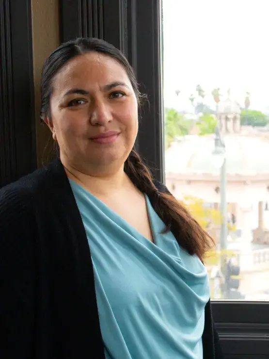 Person with long dark hair pulled into a ponytail wearing a dark jacket and light blue top with a community scene to the right