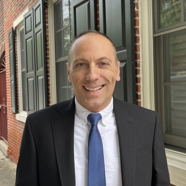 Person smiling in white collar shirt, blue tie and black jacket in front of window panes and brick building