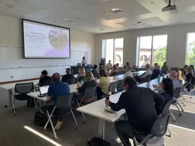Classroom setting with rows of people at long tables and a speaker and screen at the front of the room