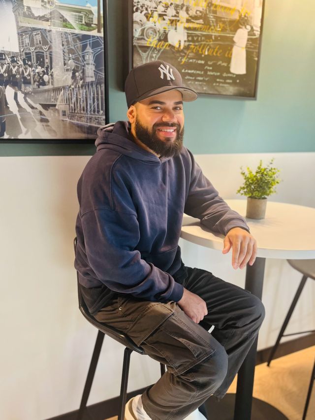 Person smiling and wearing a baseball cap sits on a stool at a high-top table