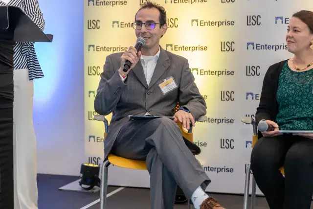 Two people seated on stage with person on left holding a mic