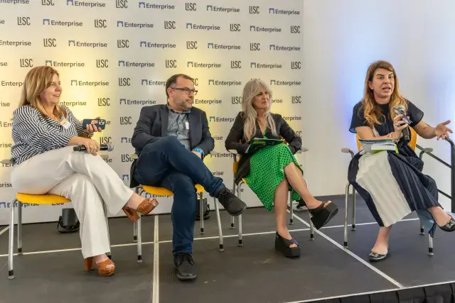 Group of four people seated on stage speaking to audience. Person on far right is holding a mic.