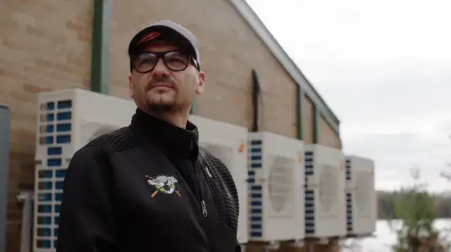 Man wearing black jacket, glasses, and hat looking into the distance in front of green housing