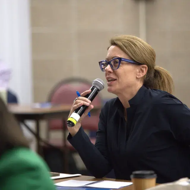 Person wearing glasses and ponytails sits at table speaking into a mic