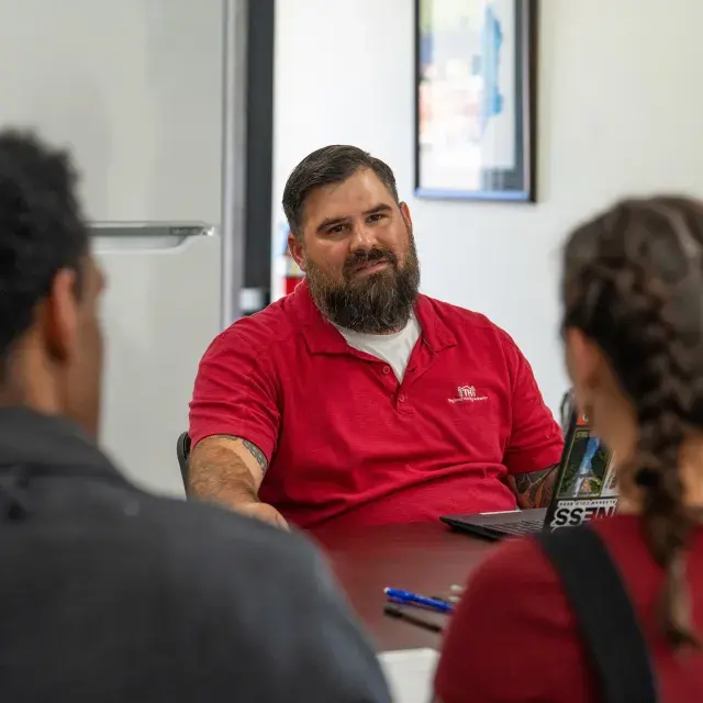 Person with dark hair and beard wearing a red shirt sits at table across from two other people