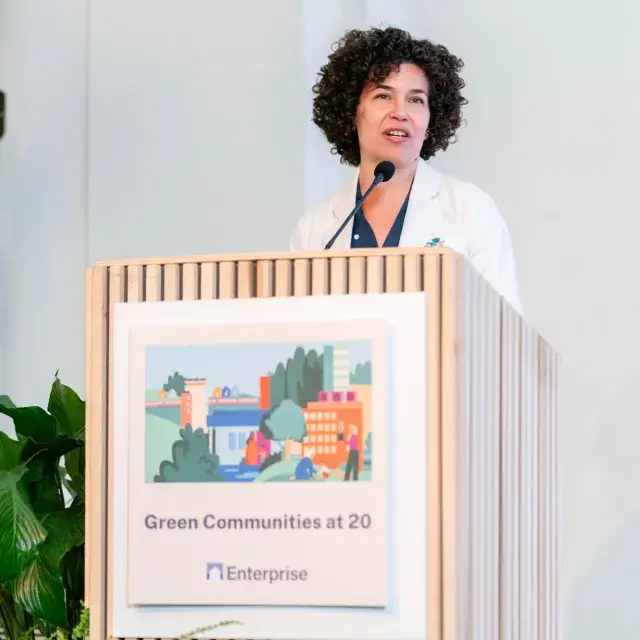 Person wearing white jacket and dark shirt stands speaking at podium with microphone