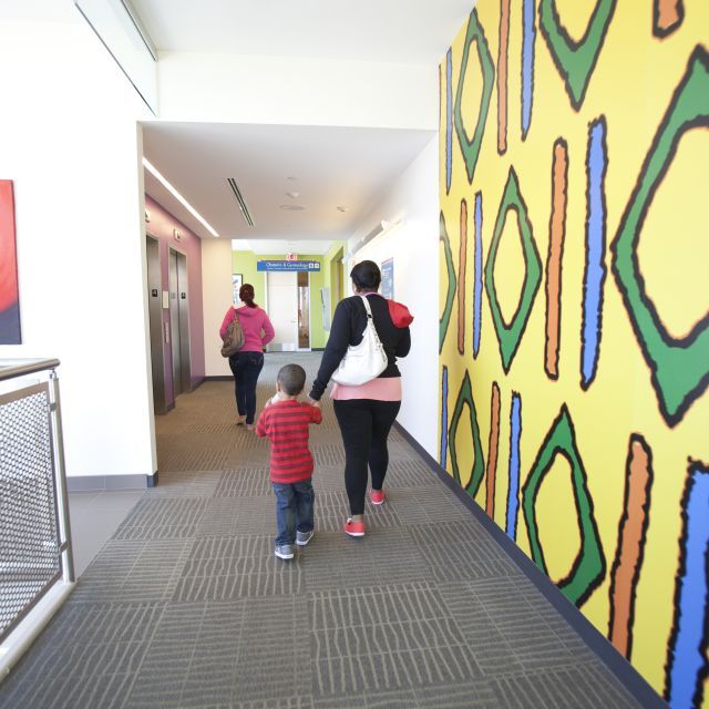 A mother and child with their backs to the camera walk down a hallway. On the right is a brightly colored wall painted with geometric shapes.