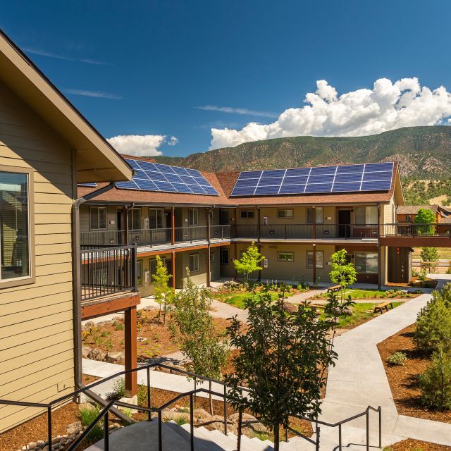 Courtyard of a multifamily housing development showing solar panels on the roof with a blue sky and white puffy clouds overhead and a mountain range in the background.