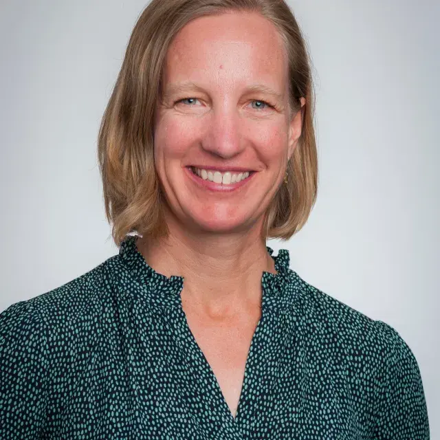 Person smiling with light brown shoulder-length hair wearing black patterned blouse.