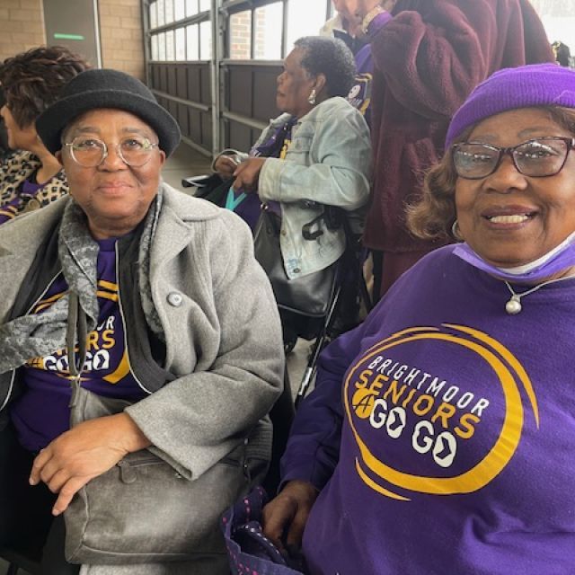 Two people sit next to each other, each wearing a purple hat, and T-shirts with the words "Brightmoor Seniors a Go Go."