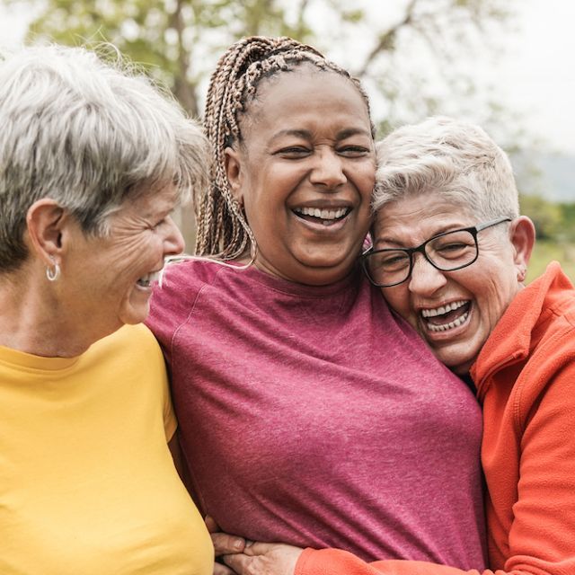 Three older women hug and laugh in front of green space.