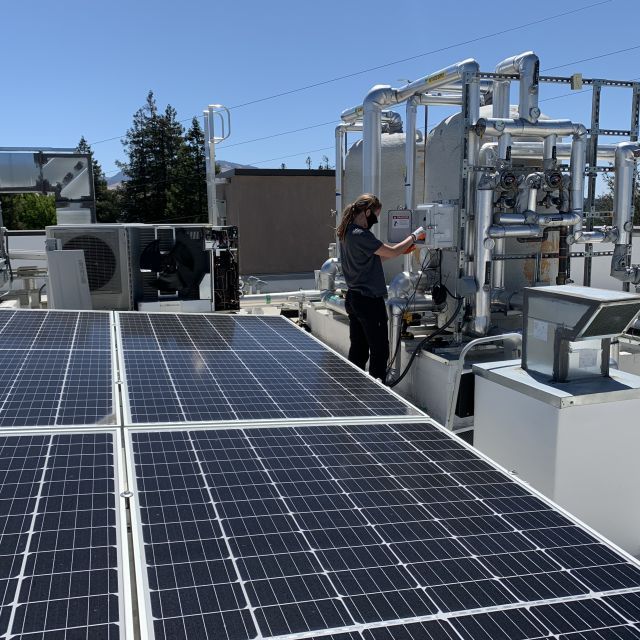 Rooftop view of flat solar panels with a worker adjusting a heat pump; blue sky and trees are in the background