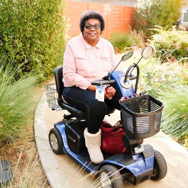 A woman on a scooter she uses as a mobility aid smiles from an apartment community's leafy green space.