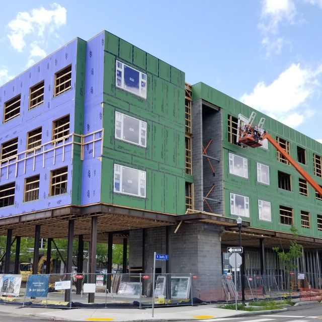 Corner multifamily apartment building undergoing a window installation; a crane is shown on the right with a blue sky and a few clouds in the background
