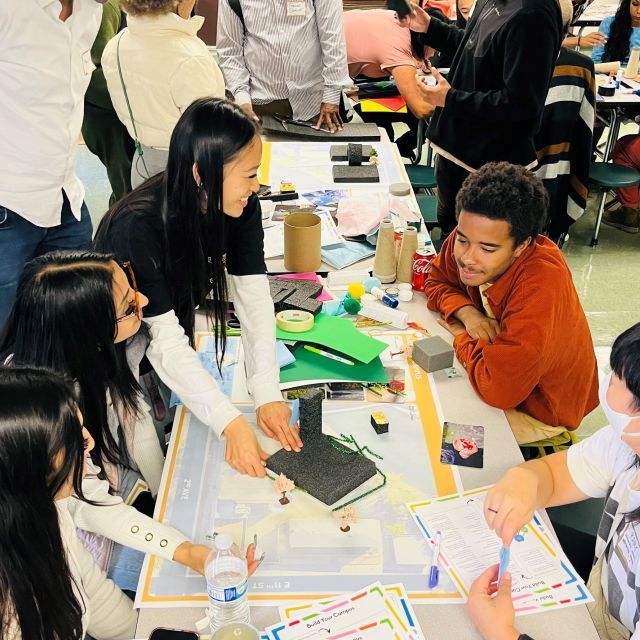 Group of smiling young people sit at a long, narrow conference table with papers and files