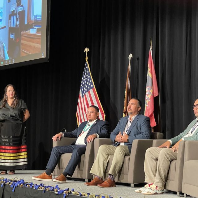 Men sitting in chairs with a woman and man standing behind a podium and a woman on a screen