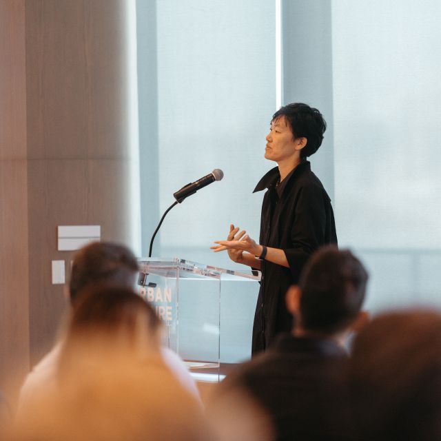 Person stands at a podium speaking into a mic before an audience of people