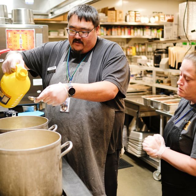 A woman looks on as a man pours liquid into two large pots on the stove with shelves and steel tables behind them