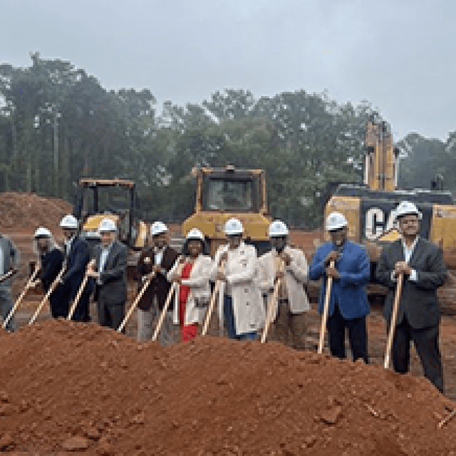 A group of people standing in a row holding shovels in the dirt with three construction vehicles behind them