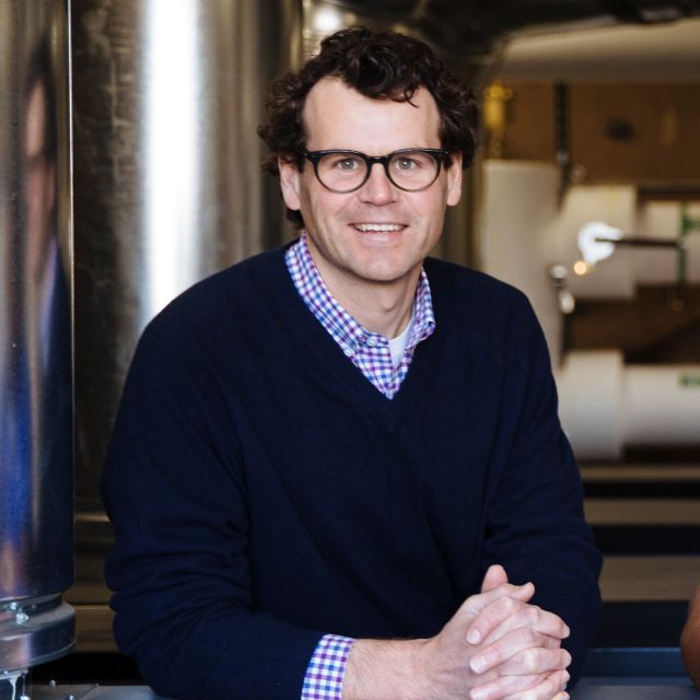 Person with glasses and short brown hair smiles wearing a black sweater and blue checkered collar shirt; hands are crossed on a table and HVAC equipment is in the background