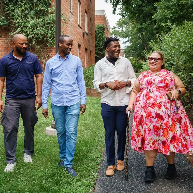 Group of four people walking down a path with trees and a brick building in the background