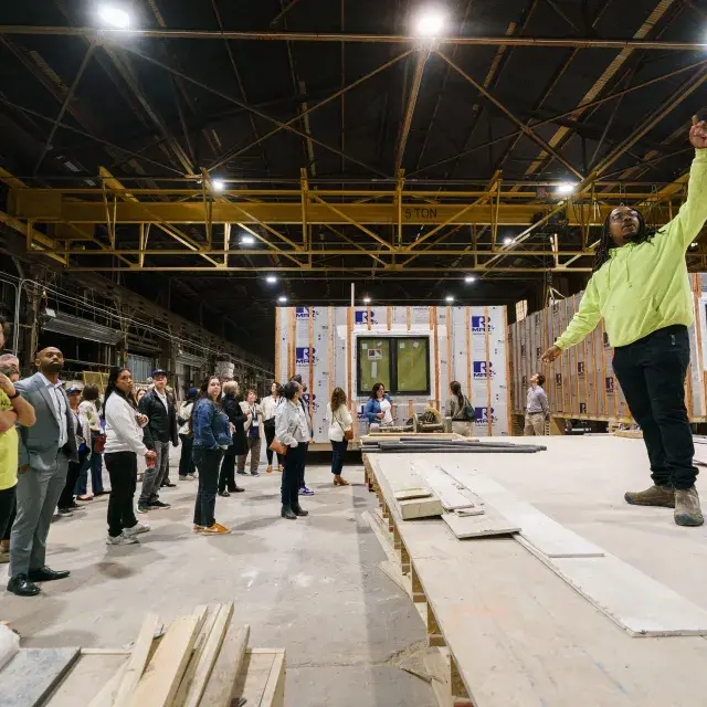 Person in neon yellow sweatshirt stands inside modular housing factory speaking to a group of people. There are modular structures in the background.
