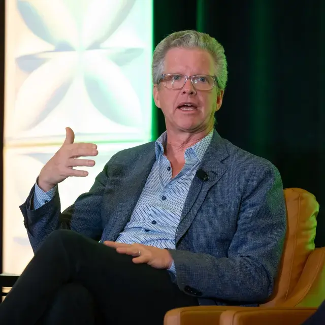 Person in blue shirt and jacket speaks, gesturing with their hand