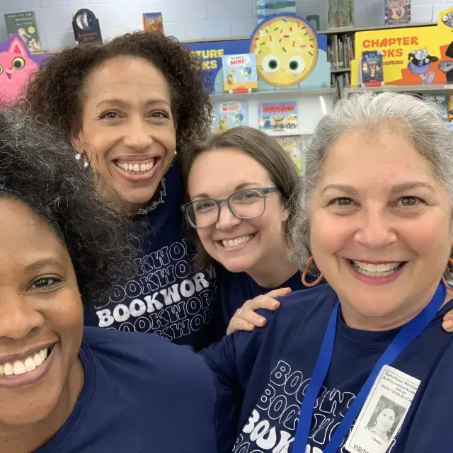 Group of four women stand next to each other smiling with children's bookcase in the background