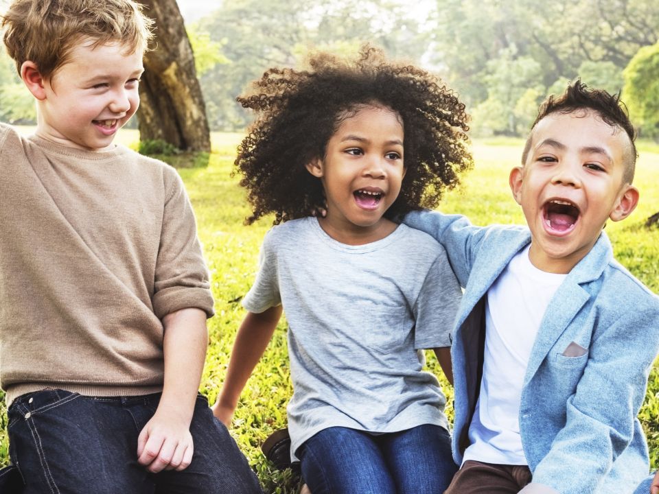 three kids of various ethnic backgrounds sitting in the grass laughing