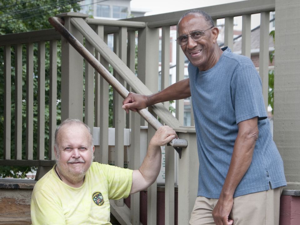 An older adult male standing next to an older adult male sitting on the steps
