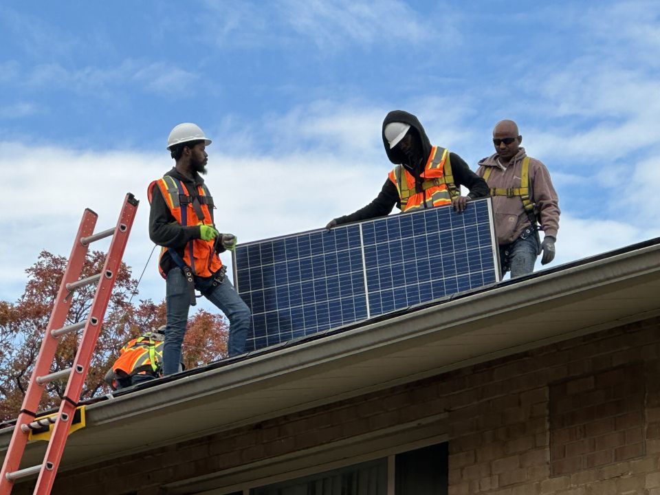 Two solar panel installers hold a solar panel during an installation on a rooftop as another installer looks on