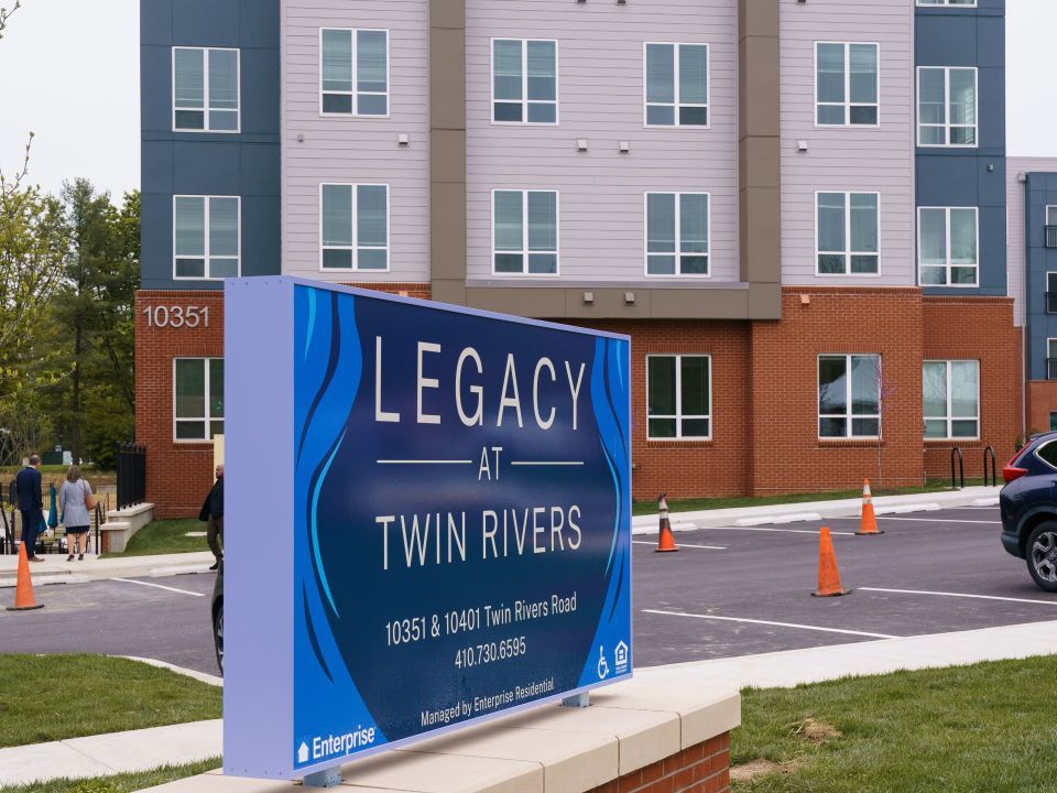 Property entrance sign for the Legacy at Twin Rivers with a parking lot and one of the apartment buildings in the background