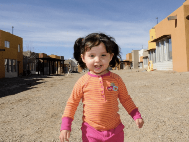 A little girl with an orange shirt smiling while standing on a construction site.