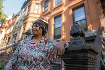 Woman stands on a sidewalk leaning on the banister of the brownstone steps